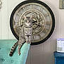 cat, gray_tabby, chair, clock, vintage_clock, indoor, furniture, wall, decor, pet, sitting, curious, calm, teal_chair, roman_numerals, timepiece, striped_cat, tail, ears, whiskers