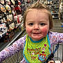 toddler, child, shopping_cart, bib, clothing, hair_tie, shopping_aisle, store, retail, face, smile, rosy_cheeks, purple_outfit, toy, colorful, indoor, person, cute, young_child, happy