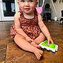 toddler, child, toy, floor, dress, patterned_clothing, barefoot, indoor, curious, smiling, sitting, baby, playtime, hands, feet, face, person, home, light, portrait