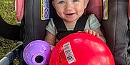 Hazel joined the competition — help win amazing prizes! baby, car_seat, red_ball, purple_toy, smiling, happy, child, outdoor, grass, backpack, plastic_bottle, clothing, seatbelt, person, toy, head, face, legs, water_bottle, sunlight