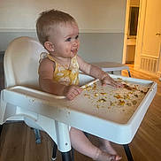 Hazel joined the competition — help win amazing prizes! baby, high_chair, messy, food, yellow_clothing, floor, wooden_floor, face, small_child, eating, home_interior, family_photos, wall, doorway, foot, hand, table, room, person, happy
