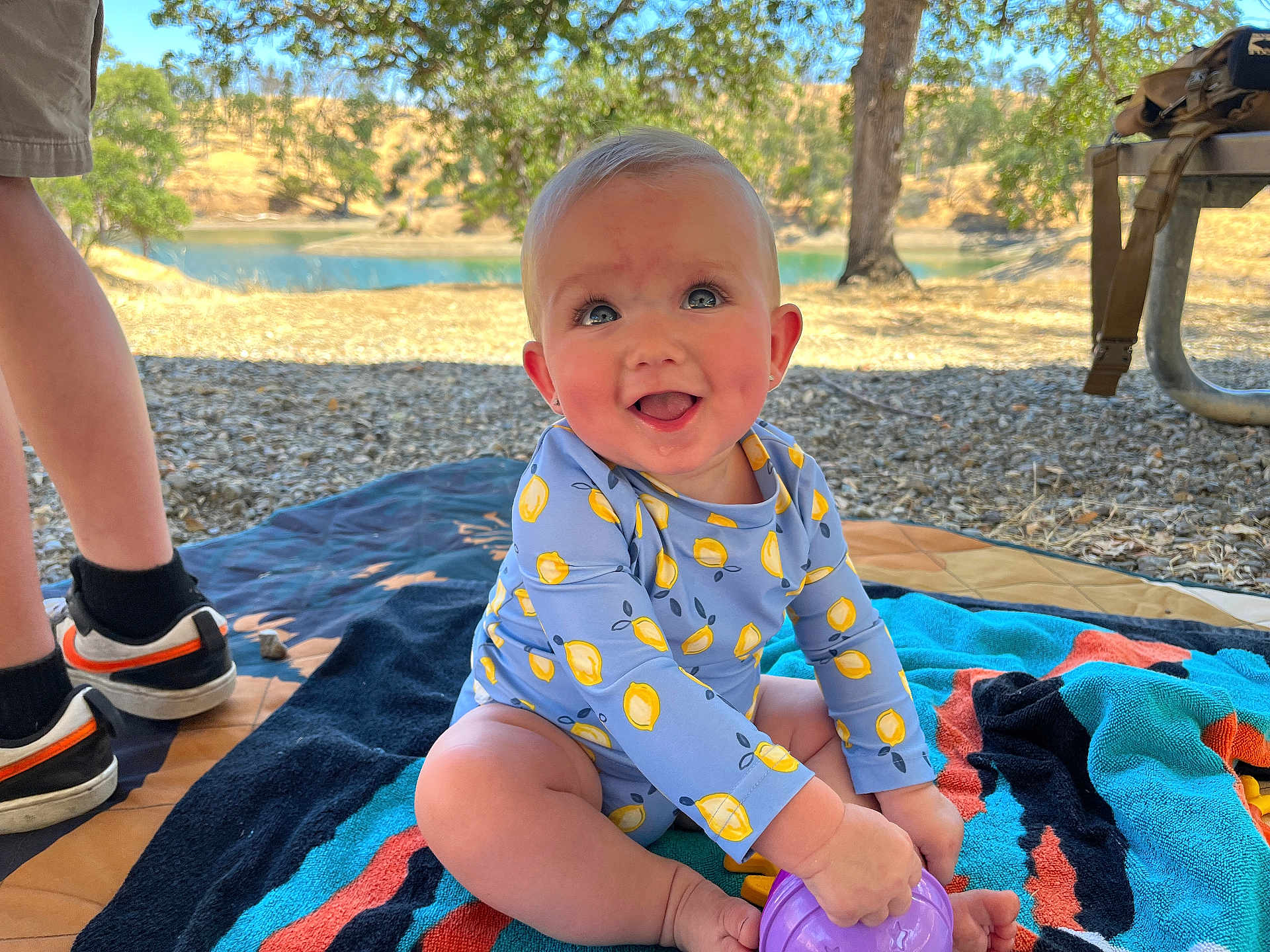Hazel is registered to the contest to win money with this photo: baby, child, outdoor, blue_eyes, smiling, toy, towel, tree, lake, sunlight, grass, shorts, sneakers, legs, nature, picnic, playing, happy, person, summer