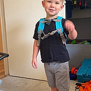 Tucker is registered to the contest to win money with this photo: child, boy, smiling, indoor, carpet, backpack, red_shoes, shorts, black_tshirt, toy, closet, clothing, carpeted_floor, door, furniture, casual, happy, standing, young_child, home