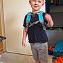 child, boy, smiling, indoor, carpet, backpack, red_shoes, shorts, black_tshirt, toy, closet, clothing, carpeted_floor, door, furniture, casual, happy, standing, young_child, home