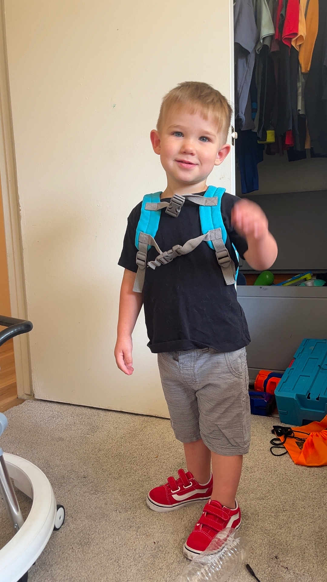 Tucker is registered to the contest to win money with this photo: child, boy, smiling, indoor, carpet, backpack, red_shoes, shorts, black_tshirt, toy, closet, clothing, carpeted_floor, door, furniture, casual, happy, standing, young_child, home