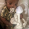 baby, pacifier, plush_toy, bear, camo_clothing, infant, child, cute, soft_toy, bed, white_bed, sleepwear, hand, face, lying_down, indoor, resting, comfort, toddler, person