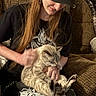 Beanie is registered to the contest to win money with this photo: person, cat, indoor, couch, long_hair, piercing, black_cap, black_tshirt, pet, lap, brown_couch, casual_clothing, relaxed, affection, seated, feline, human, warm_lighting, textured_fabric, home