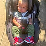 baby, car_seat, child, sneakers, green_pants, sunlight, outdoor, concrete, seat_belt, infant, clothing, footwear, sitting, person, casual, daylight, shadow, safety, cute, portrait