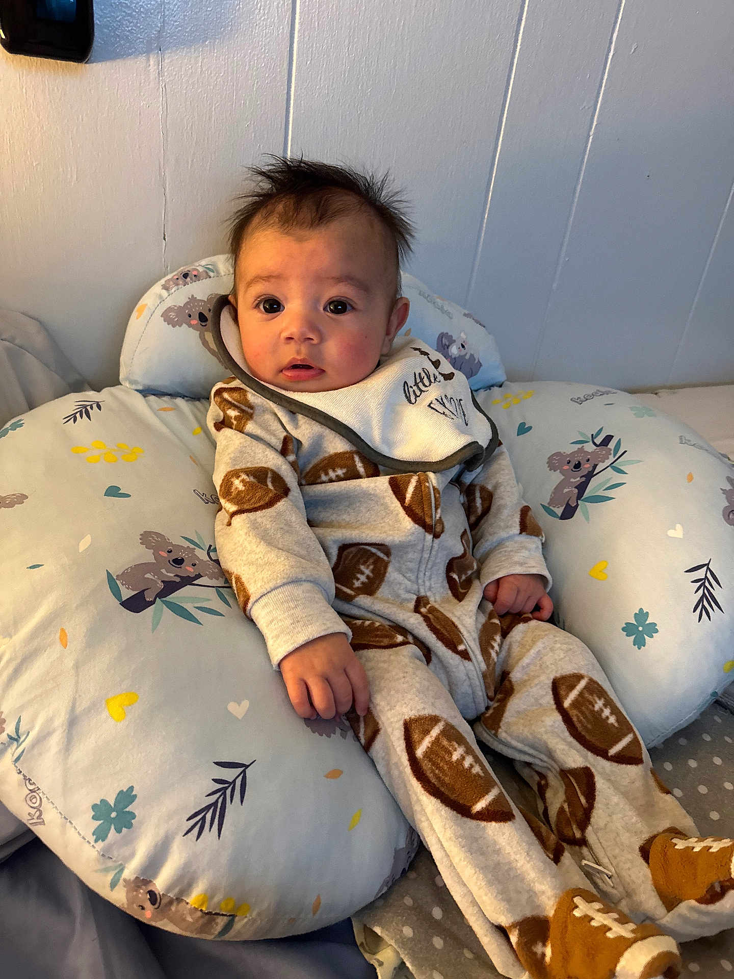 Samuel is registered to the contest to win money with this photo: baby, infant, onesie, football_pattern, pillow, koala_pattern, indoor, cute, child, face, expression, clothing, footwear, sitting, soft_furnishing, comfort, bedroom, young_child, portrait, casual