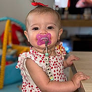 Ambre a rejoint le concours — aidez-le/la à gagner de superbes lots ! baby, child, pacifier, red_bow, cute, indoor, table, clothing, face, person, smiling, beads, toy, hand, arm, skin, young_child, portrait, focus, background_blur