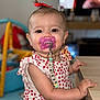 baby, child, pacifier, red_bow, cute, indoor, table, clothing, face, person, smiling, beads, toy, hand, arm, skin, young_child, portrait, focus, background_blur