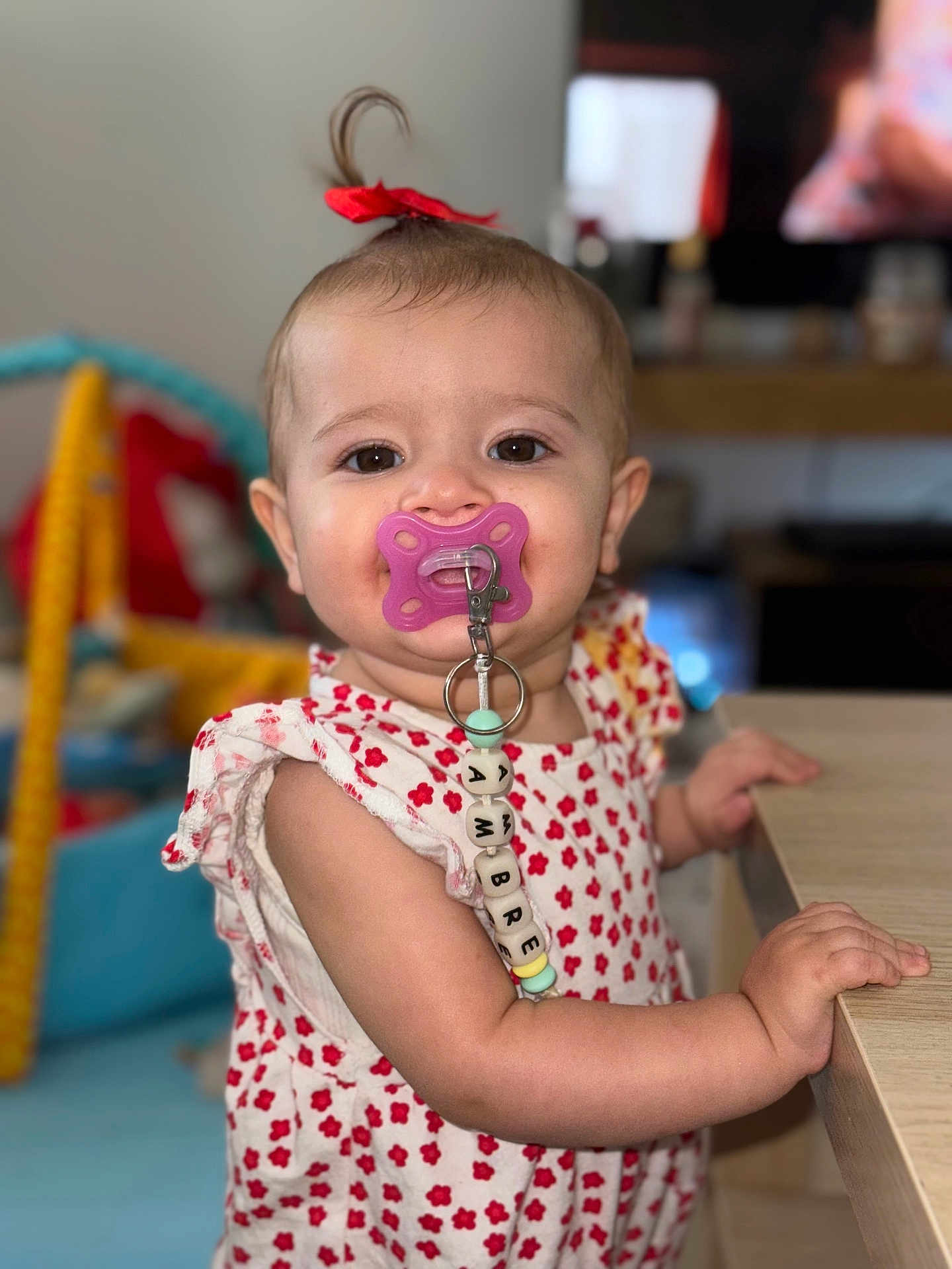 Ambre a rejoint le concours — aidez-le/la à gagner de superbes lots ! baby, child, pacifier, red_bow, cute, indoor, table, clothing, face, person, smiling, beads, toy, hand, arm, skin, young_child, portrait, focus, background_blur