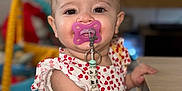 Ambre a rejoint le concours — aidez-le/la à gagner de superbes lots ! baby, child, pacifier, red_bow, cute, indoor, table, clothing, face, person, smiling, beads, toy, hand, arm, skin, young_child, portrait, focus, background_blur