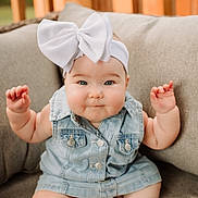Stella is registered to the contest to win money with this photo: baby, child, denim_vest, white_bow, couch, cushion, indoor, smile, cute, portrait, sitting, face, skin, happy, infant, headwear, person, plush, soft_lighting, closeup