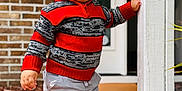 Kylian is registered to the contest to win money with this photo: toddler, child, sweater, beanie, pants, shoes, white_shoes, red_sweater, striped_sweater, porch, brick_wall, door, standing, hand_on_post, outdoor, casual_clothing, young_child, cute, baby_face, winter_clothing