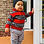 toddler, child, sweater, beanie, pants, shoes, white_shoes, red_sweater, striped_sweater, porch, brick_wall, door, standing, hand_on_post, outdoor, casual_clothing, young_child, cute, baby_face, winter_clothing