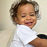 baby, casual_clothing, child, curly_hair, cushion, cute, face, happy, indoor, light_background, mickey_mouse, person, playful, portrait, relaxed, sitting, smiling, toddler, white_tshirt, young_child