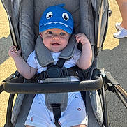 Reggie is registered to the contest to win money with this photo: baby, baby_boy, baby_clothes, blue_hat, child, footwear, hand, happy, hat, infant, legs, outdoor, pavement, person, seat_belt, shadow, smile, stroller, sunlight, white_clothes