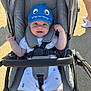 baby, baby_boy, baby_clothes, blue_hat, child, footwear, hand, happy, hat, infant, legs, outdoor, pavement, person, seat_belt, shadow, smile, stroller, sunlight, white_clothes