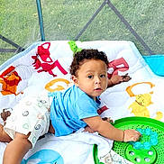 Ishmael joined the competition — help win amazing prizes! baby, child, curly_hair, blue_shirt, white_shorts, smiley_faces, green_toy, play_mat, indoor, mesh_screen, grass, cute, infant, lying_down, toddler, colorful, playful, person, young_child, curious