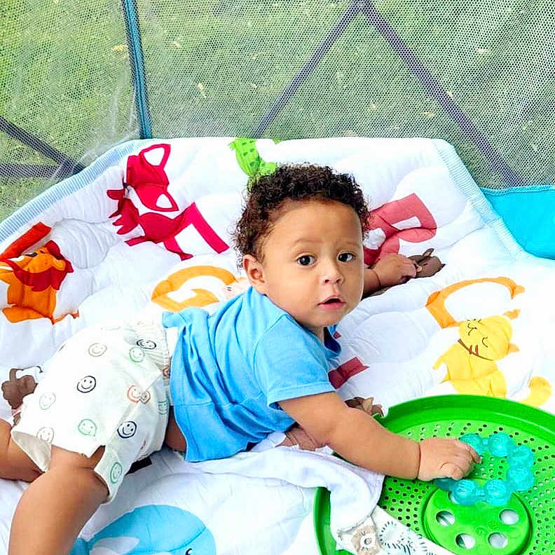 Ishmael joined the competition — help win amazing prizes! baby, child, curly_hair, blue_shirt, white_shorts, smiley_faces, green_toy, play_mat, indoor, mesh_screen, grass, cute, infant, lying_down, toddler, colorful, playful, person, young_child, curious
