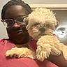 Elle is registered to the contest to win money with this photo: dog, puppy, fluffy, cream_colored, pet, portrait, selfie, woman, person, eyeglasses, indoor, home, holding, cuddling, paws, fur, pink_shirt, smiling, companion, closeup