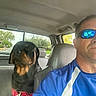 man, dog, rottweiler, car_interior, sunglasses, seatbelt, driver, pet, vehicle, road, window, daytime, casual_clothing, reflection, backseat, leather_seat, portrait, outdoor, serious_expression, animal