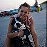 woman, dog, outdoor, pavement, hugging, black_and_white, glasses, smiling, casual_clothing, sunset, people, car, blurred_background, kneeling, friendship, pet, happy, candid, animal, leisure