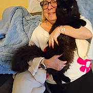 Artemis participe au concours pour gagner de l'argent avec cette photo : woman, glasses, cat, black_cat, fluffy_cat, pet, couch, blanket, plush_toy, white_sweater, pink_flowers, smiling, indoor, cozy, watch, bracelet, happy, holding, seated, furniture