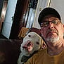 man, dog, glasses, cap, indoor, couch, brown, white, beard, facial_hair, seated, relaxed, portrait, casual, home, furniture, person, pet, companion, closeup
