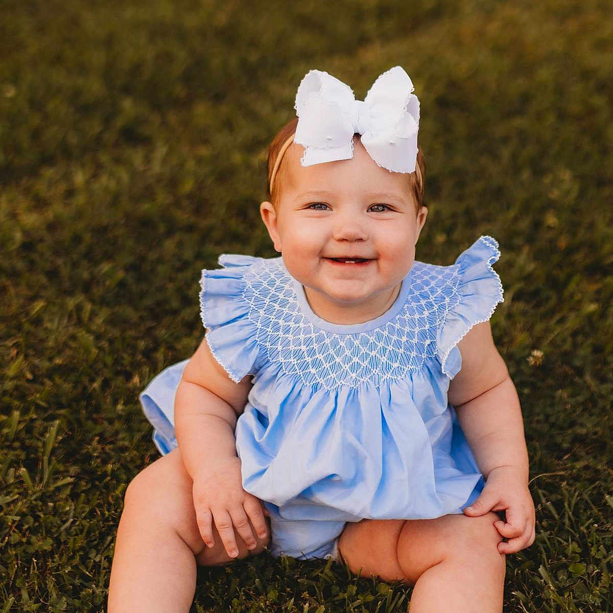 Lainey Beth is registered to the contest to win money with this photo: baby, baby_clothing, blue_dress, child, cute, face, grass, greenery, happy, headband, infant, nature, one_person, outdoor, person, portrait, sitting, smiling, sunlight, white_bow
