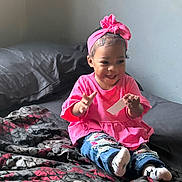 Zyura is registered to the contest to win money with this photo: bed, blanket, casual_clothing, child, clapping, cute, happy, headband, home, indoors, patterned_blanket, person, pink_clothing, playful, portrait, seated, smiling, socks, toddler, young_child