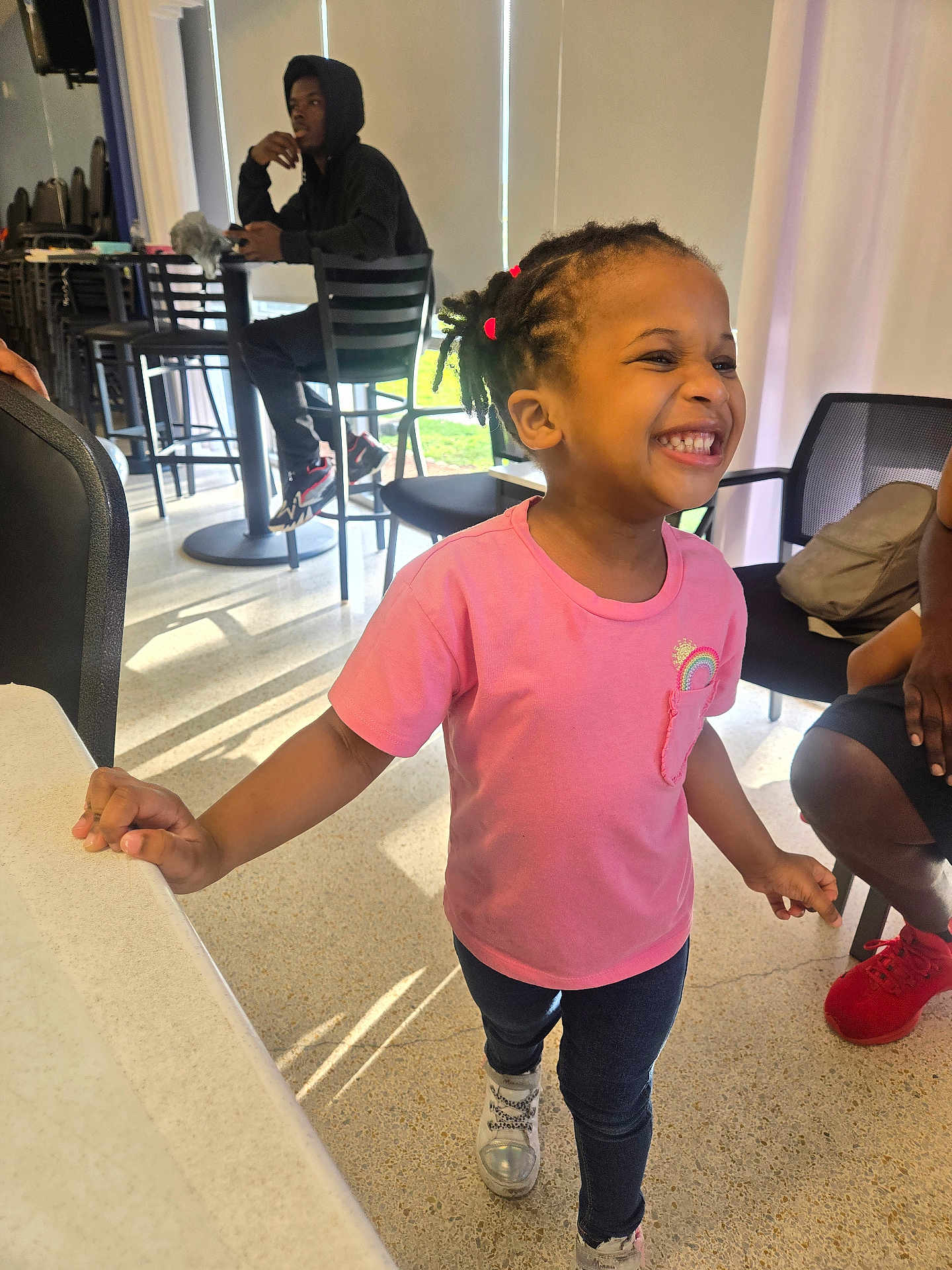 Talea is registered to the contest to win money with this photo: child, girl, smiling, pink_shirt, jeans, sneakers, indoor, table, chair, person, happy, hair_ties, dreadlocks, casual, footwear, young_adult, hoodie, seated, daylight, floor