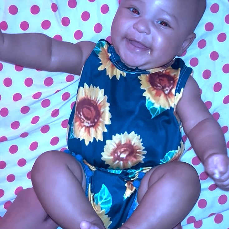 Ariarae is registered to the contest to win money with this photo: arms, baby, blanket, child, colorful, cute, face, feet, happy, indoors, infant, lying_down, patterned_clothing, person, polka_dot, portrait, skin, smiling, sunflower, toddler