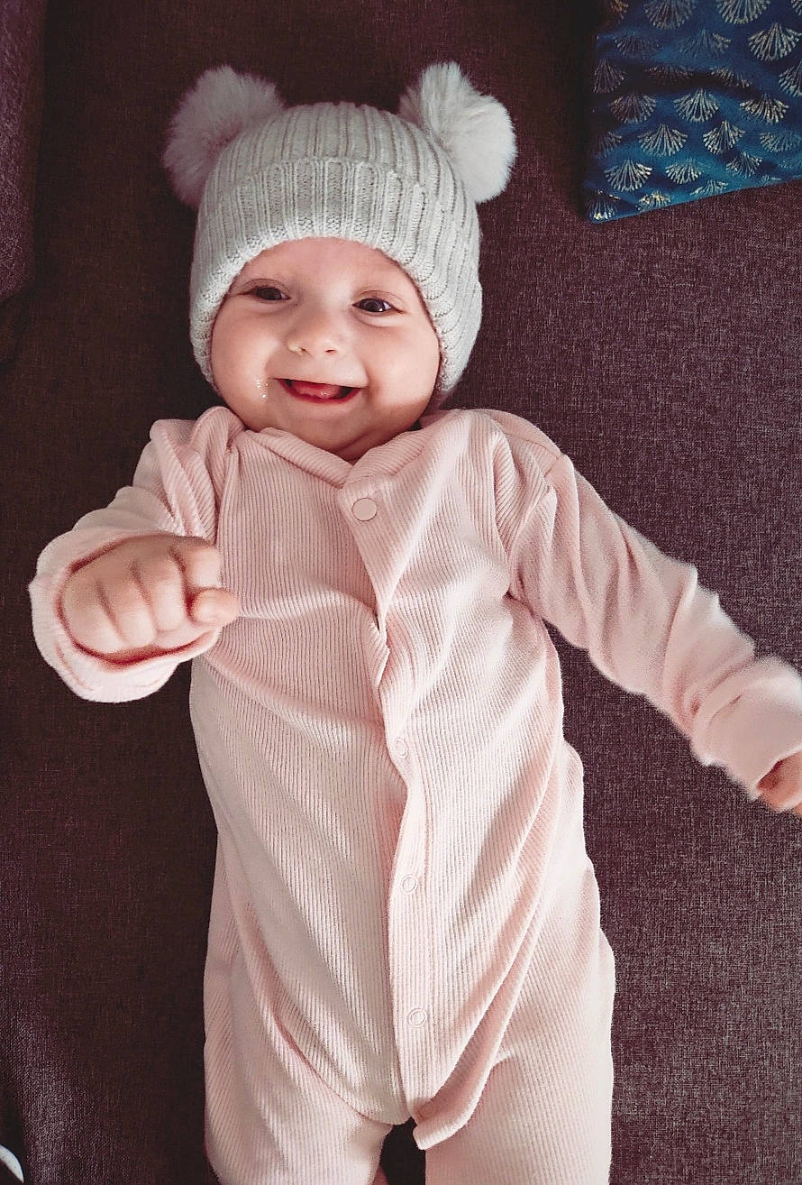 Anaë participe au concours pour gagner de l'argent avec cette photo : baby, baby_toddler_clothing, cap, collar, eye, fashion_accessory, gesture, happy, headgear, headwear, joy, knit_cap, magenta, outerwear, pattern, person, pink, sleeve, smile, thumb