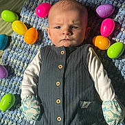 Joshua is registered to the contest to win money with this photo: baby, infant, child, blanket, crochet_blanket, easter_eggs, plastic_eggs, mittens, onesie, buttons, portrait, indoor, toy, colorful, blue, pink, yellow, green, sunlight, expression_frown