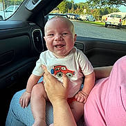 Camden is registered to the contest to win money with this photo: baby, smiling, child, person, car_interior, window, hand, clothing, shirt, embroidery, red_car, lap, happy, outdoor_view, trees, parking_lot, vehicles, daylight, casual, cute