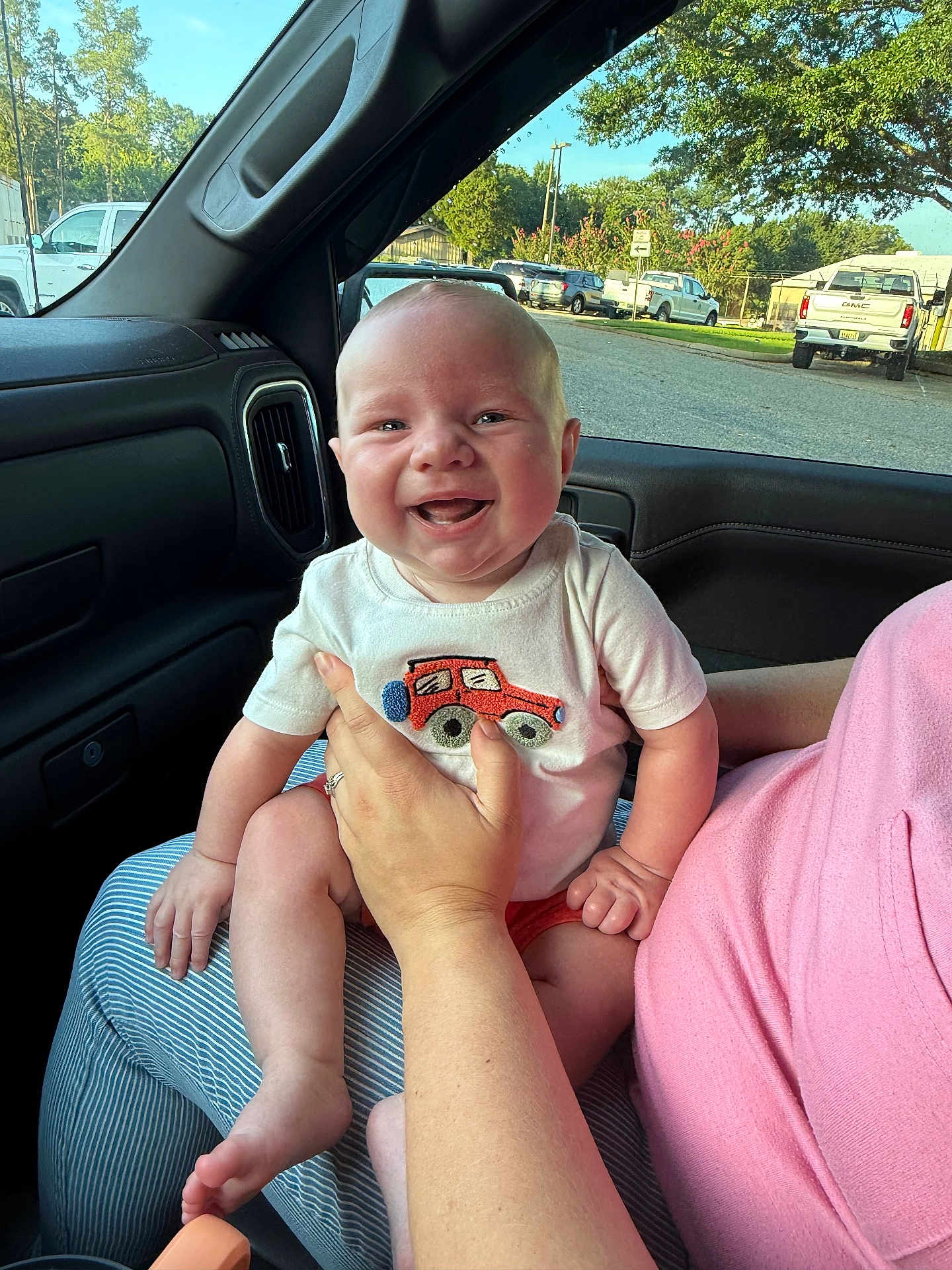 Camden is registered to the contest to win money with this photo: baby, smiling, child, person, car_interior, window, hand, clothing, shirt, embroidery, red_car, lap, happy, outdoor_view, trees, parking_lot, vehicles, daylight, casual, cute