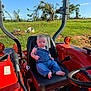 Camden is registered to the contest to win money with this photo: baby, denim_overalls, tractor, seat, outdoor, grass, sky, greenery, child, person, sunlight, farm, vehicle, steering_wheel, footwearless, serious_expression, nature, daytime, agriculture, rural