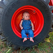Camden is registered to the contest to win money with this photo: baby, denim_overalls, tractor_tire, red_hub, outdoor, grass, dirt, baby_boy, child, face, barefoot, nature, equipment, farm, curious, sitting, small_child, clothing, person, closeup