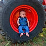 baby, denim_overalls, tractor_tire, red_hub, outdoor, grass, dirt, baby_boy, child, face, barefoot, nature, equipment, farm, curious, sitting, small_child, clothing, person, closeup