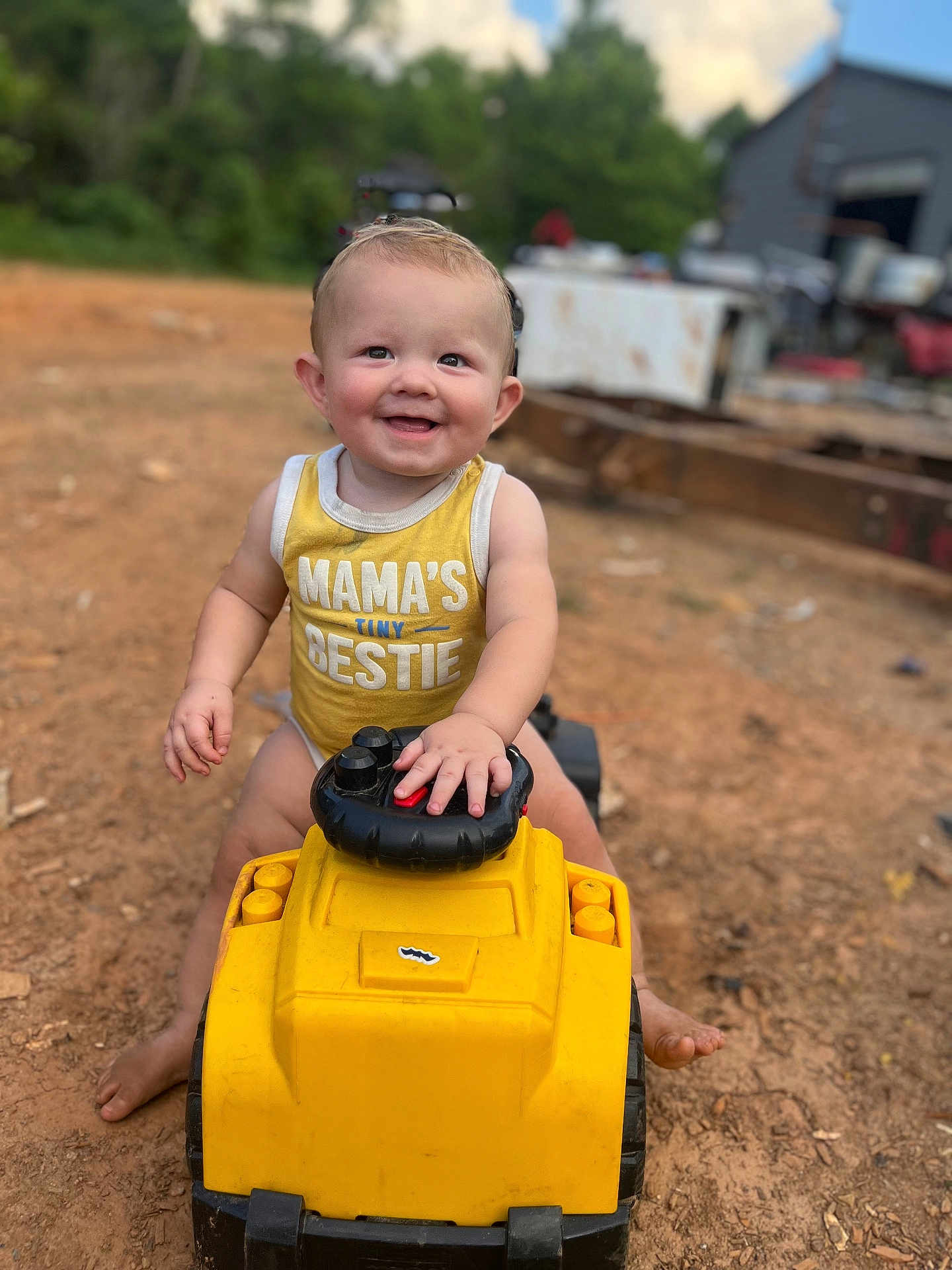 Easton is registered to the contest to win money with this photo: toddler, child, smiling, yellow_clothing, toy_vehicle, outdoor, dirt, happy, cute, person, summer, play, barefoot, tank_top, baby, nature, greenery, daylight, fun, portrait