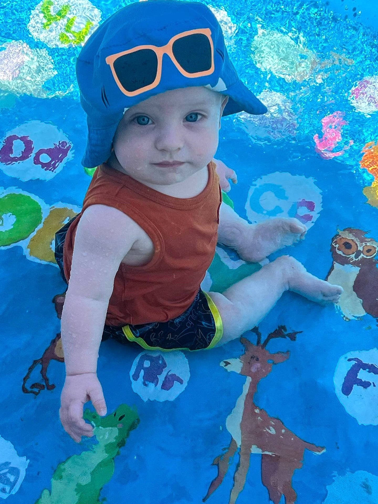 animal_prints, baby, blue_hat, child, colorful, cute, eyes, face, hand, letters, orange_tank_top, outdoor, playful, sitting, skin, summer, toddler, toy, water, water_play_mat
