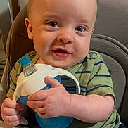 River is registered to the contest to win money with this photo: baby, child, high_chair, sippy_cup, green_shirt, striped_clothing, happy, smiling, indoor, curly_hair, face, hands, cute, infant, person, portrait, young_child, feeding, seat, expression
