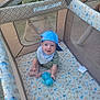 baby, bib, blue_cap, child, clothing, cute, face, grass, happy, headwear, infant, mesh, outdoor, pattern, person, playpen, sippy_cup, sitting, smiling, toy