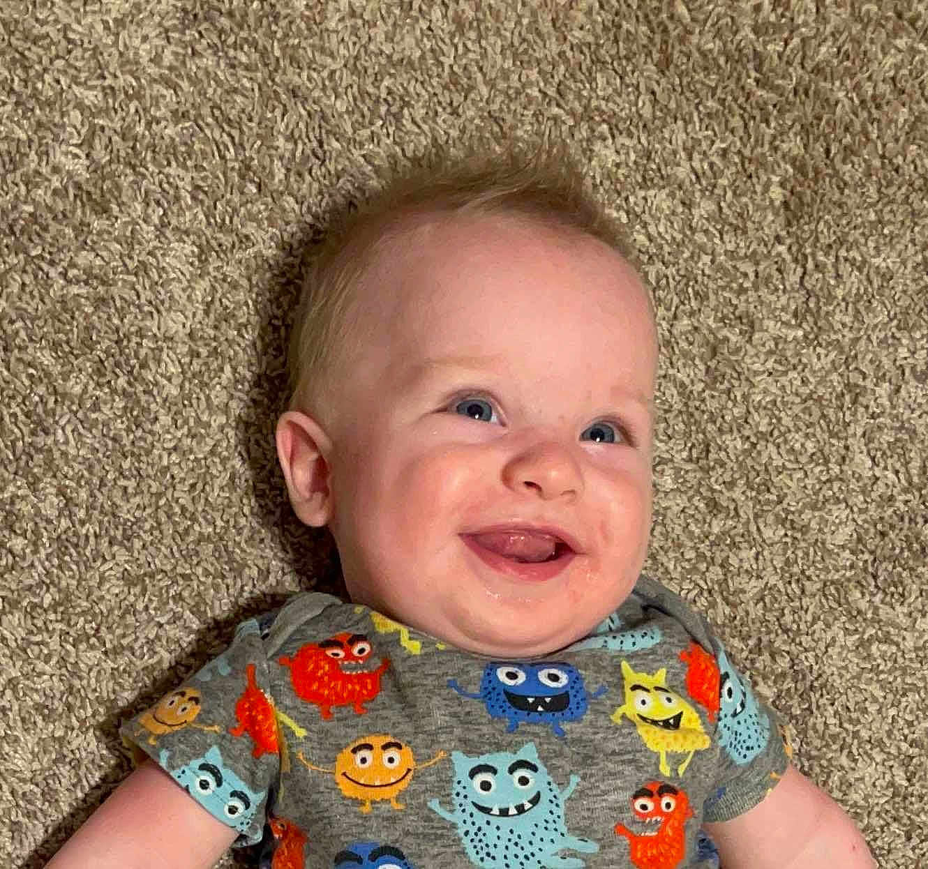 River is registered to the contest to win money with this photo: baby, infant, smile, face, blue_eyes, carpet, tshirt, cartoon_monsters, happy, child, cute, skin, short_sleeves, lying_down, indoors, expression, young, person, playful, colorful