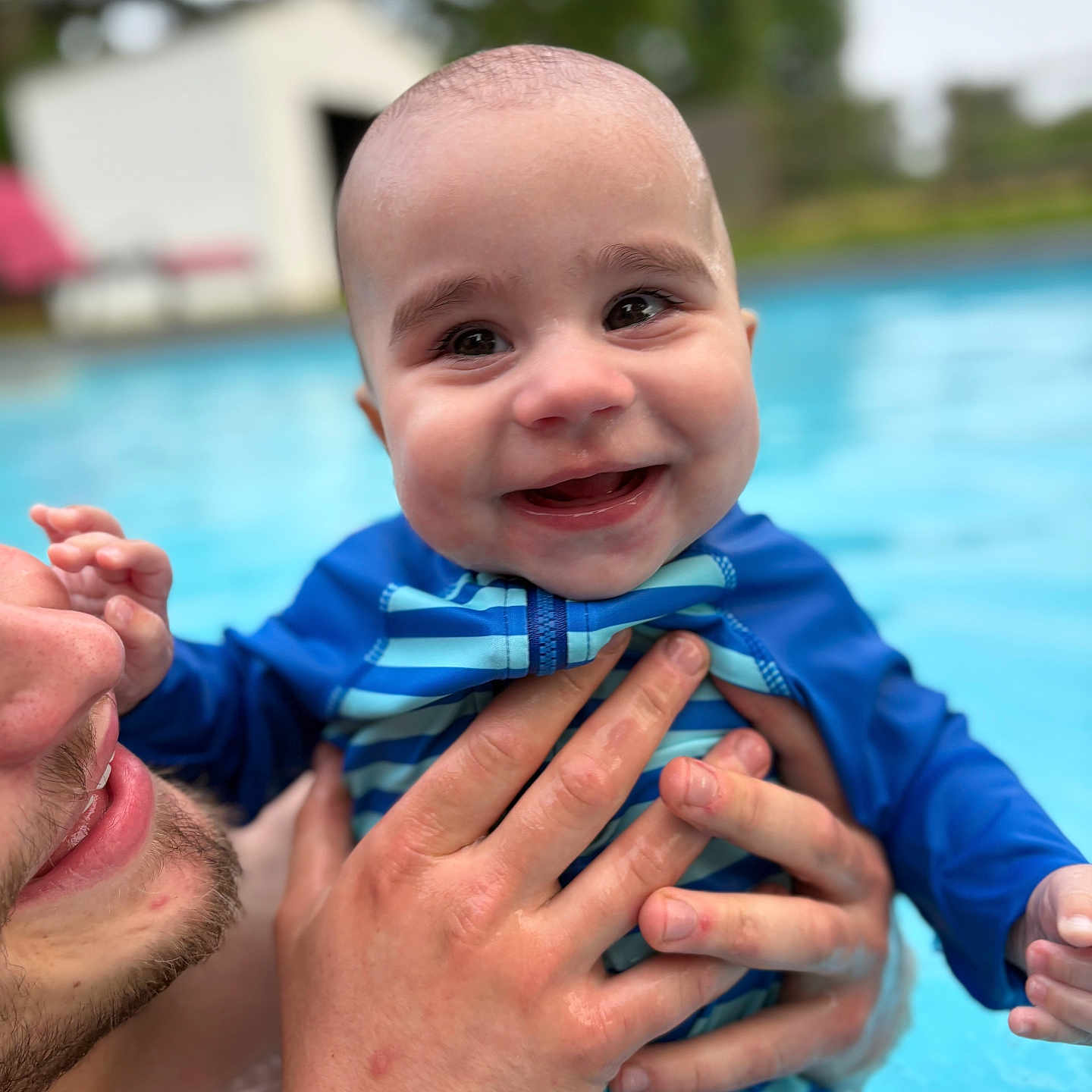 Elijah is registered to the contest to win money with this photo: adult_hand, baby, child, closeup, face, happy, head, holding, outdoor, parent, person, playful, pool, recreation, skin, smiling, striped_clothing, summer, swimsuit, water