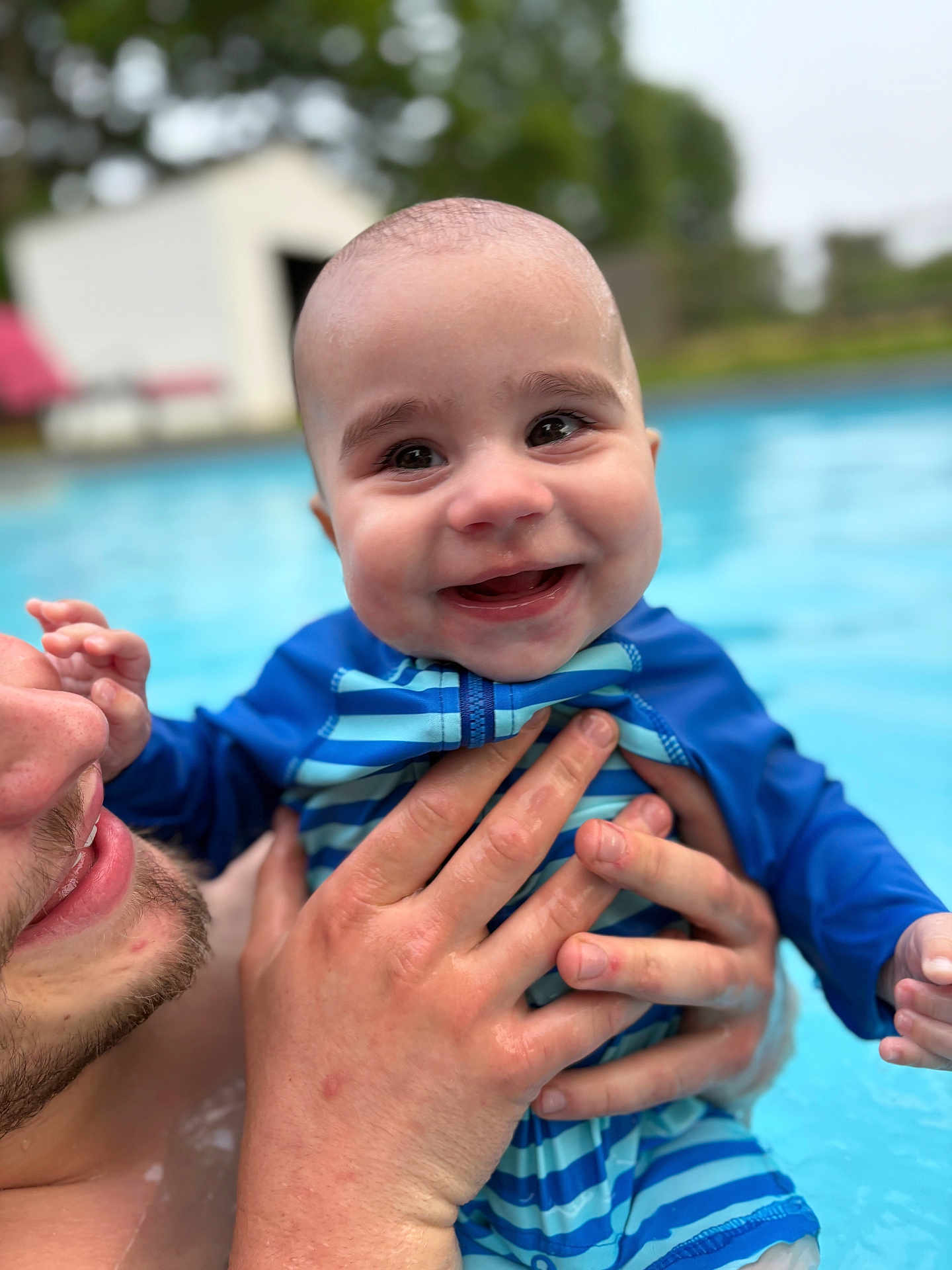 Elijah is registered to the contest to win money with this photo: baby, child, smiling, swimsuit, striped_clothing, water, pool, adult_hand, outdoor, happy, face, skin, holding, closeup, summer, recreation, playful, parent, head, person