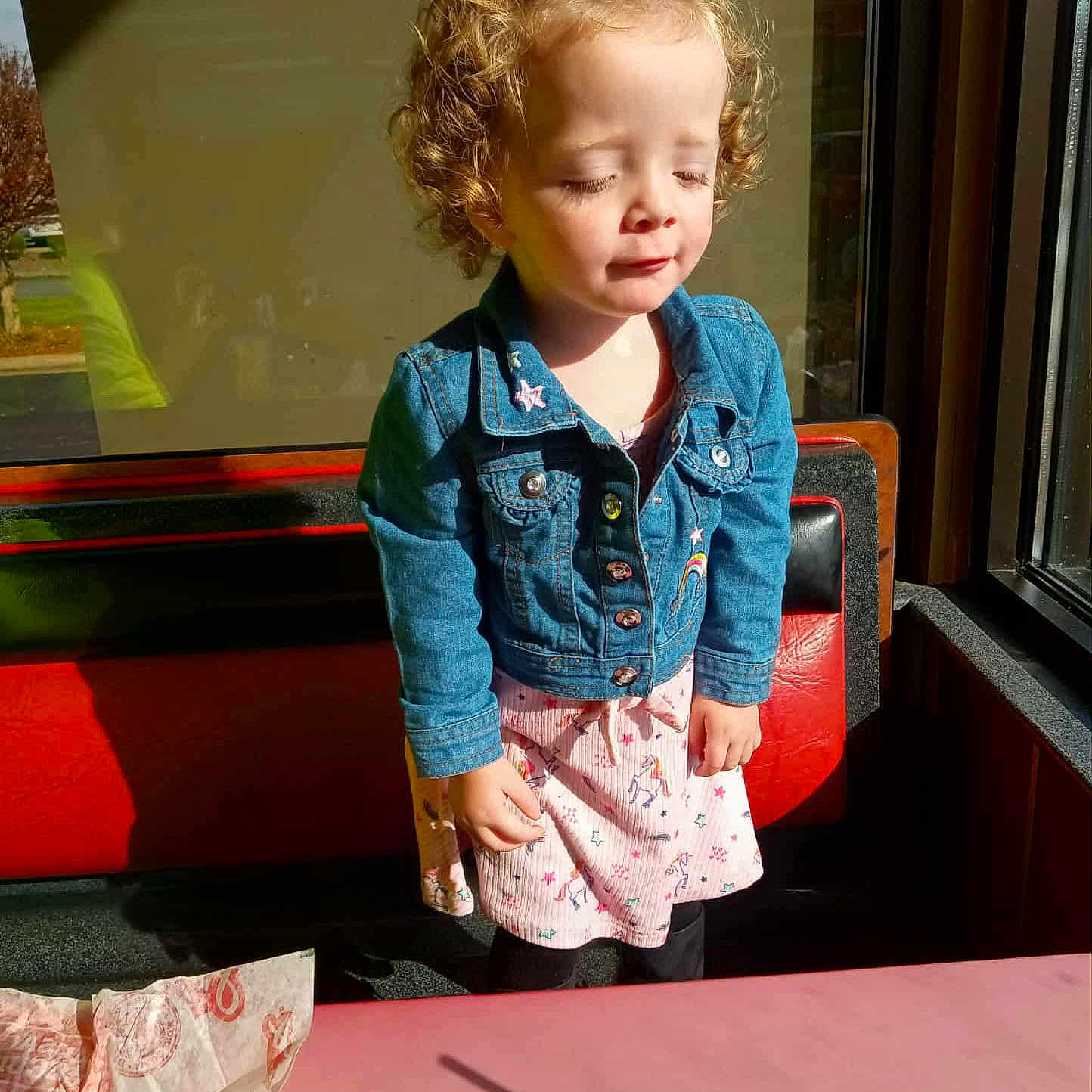 Sydney joined the competition — help win amazing prizes! bench, casual_clothing, child, closed_eyes, curly_hair, cute, daylight, denim_jacket, dress, happy, indoor, pink_table, portrait, restaurant, shadow, sunlight, table, toddler, unicorn_pattern, window