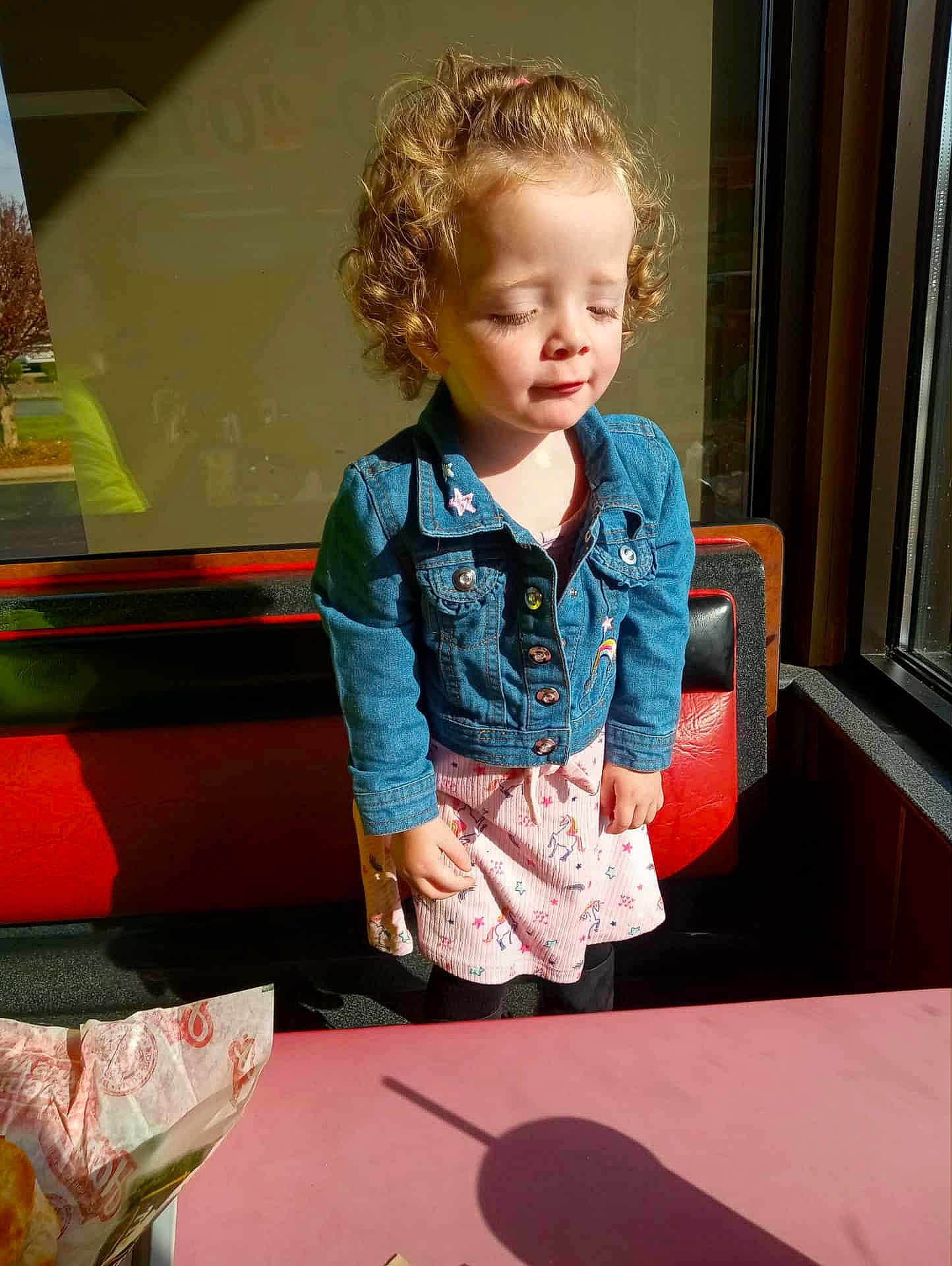 Sydney joined the competition — help win amazing prizes! child, toddler, curly_hair, denim_jacket, dress, unicorn_pattern, restaurant, bench, sunlight, shadow, window, table, pink_table, closed_eyes, cute, indoor, daylight, casual_clothing, happy, portrait